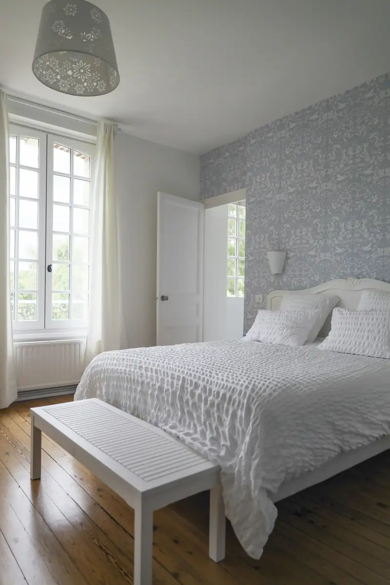 Chambre claire et terminée : lit en lin blanc, fenêtres hautes, murs fraîchement repris